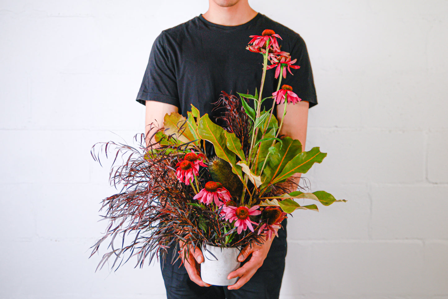 A man wearing black clothes standing against a white background holding an arrangement of pink flowers in a small, white vase