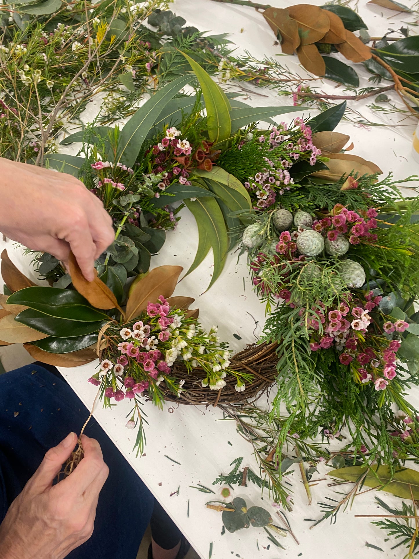 Person crafting a floral wreath with greenery and pink flowers on a white surface.