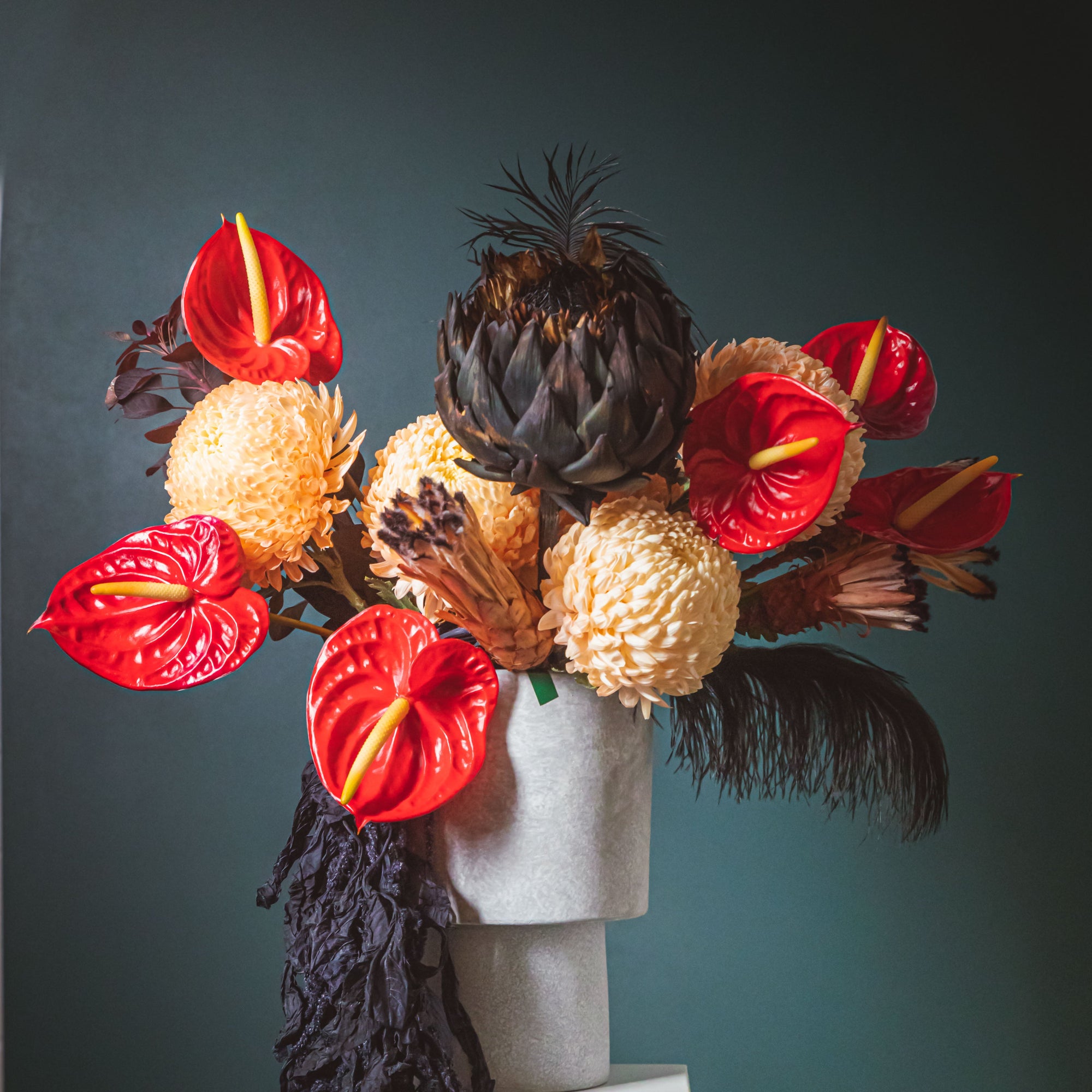 Floral arrangement with red, yellow, and black flowers in a vase on a white stool against a dark green wall.
