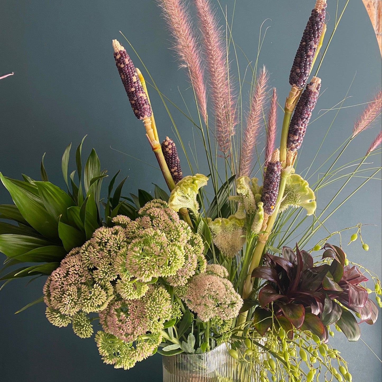 textural green and plum coloured florals in a glass w=vase with deep green backdrop