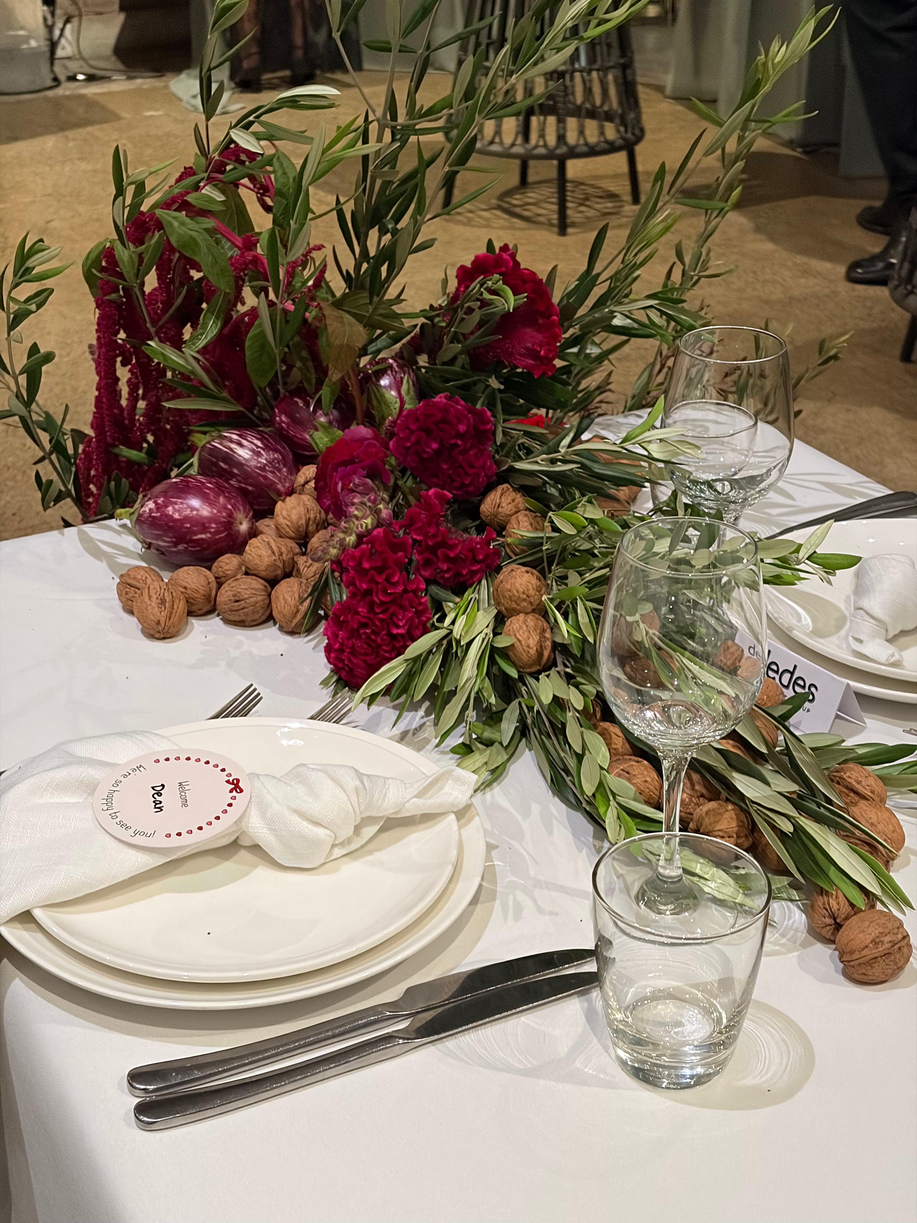 A styled dining table setting with a white tablecloth, white plates, silver cutlery, clear wine glasses, and a rustic floral centerpiece featuring deep red flowers, green olive branches, walnuts, and purple eggplants.