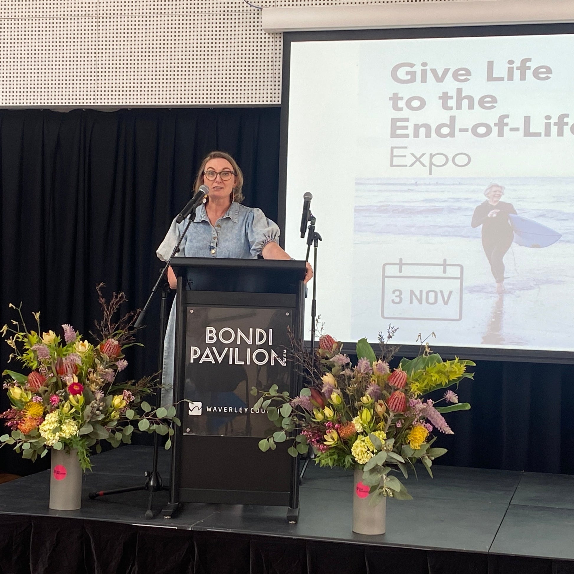 Speaker at a podium giving a talk at the "Give Life to the End-of-Life" expo with colourful flower arrangements on stage