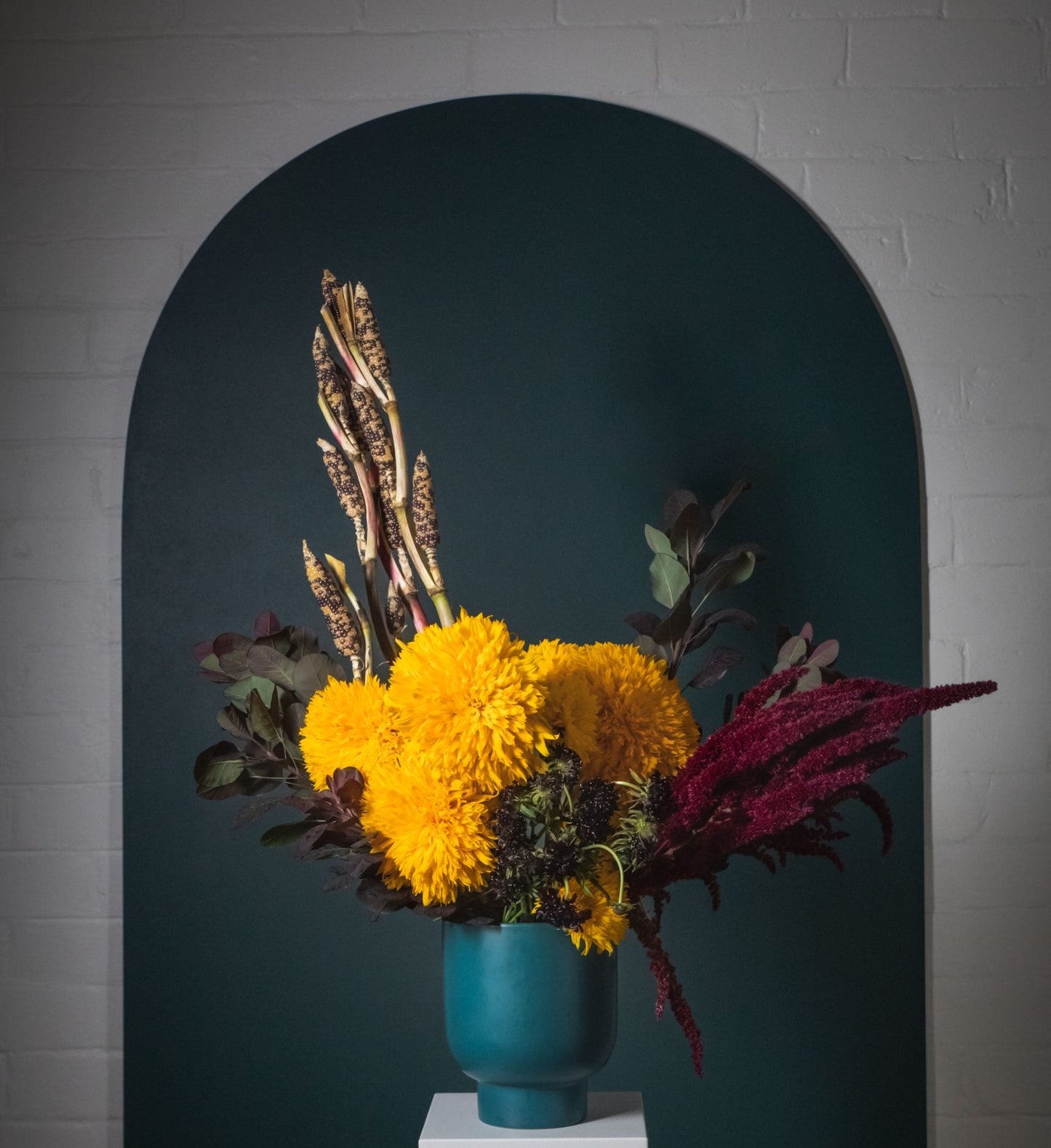 A yellow sunflower, red amarantus, smokebush and corn flower arrangement in a green vase.