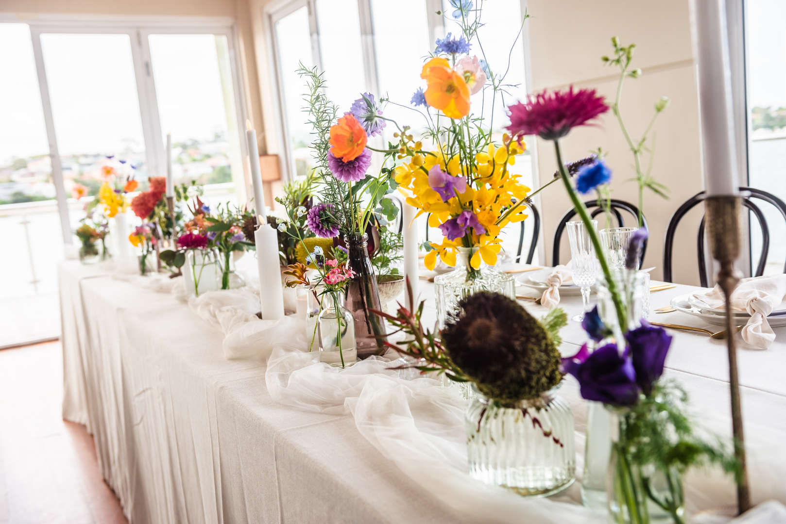A long table with white tablecloths displaying table flower arrangements of bright flowers arranged by Bold Botanicals