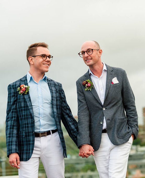 Two men holding hands with brightly coloured buttonholes made with native flowers