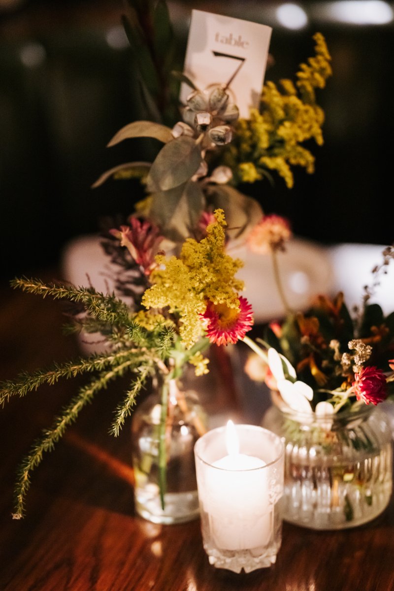 A small table arrangement of native flowers in a glass vase in a dimly lit room with candles