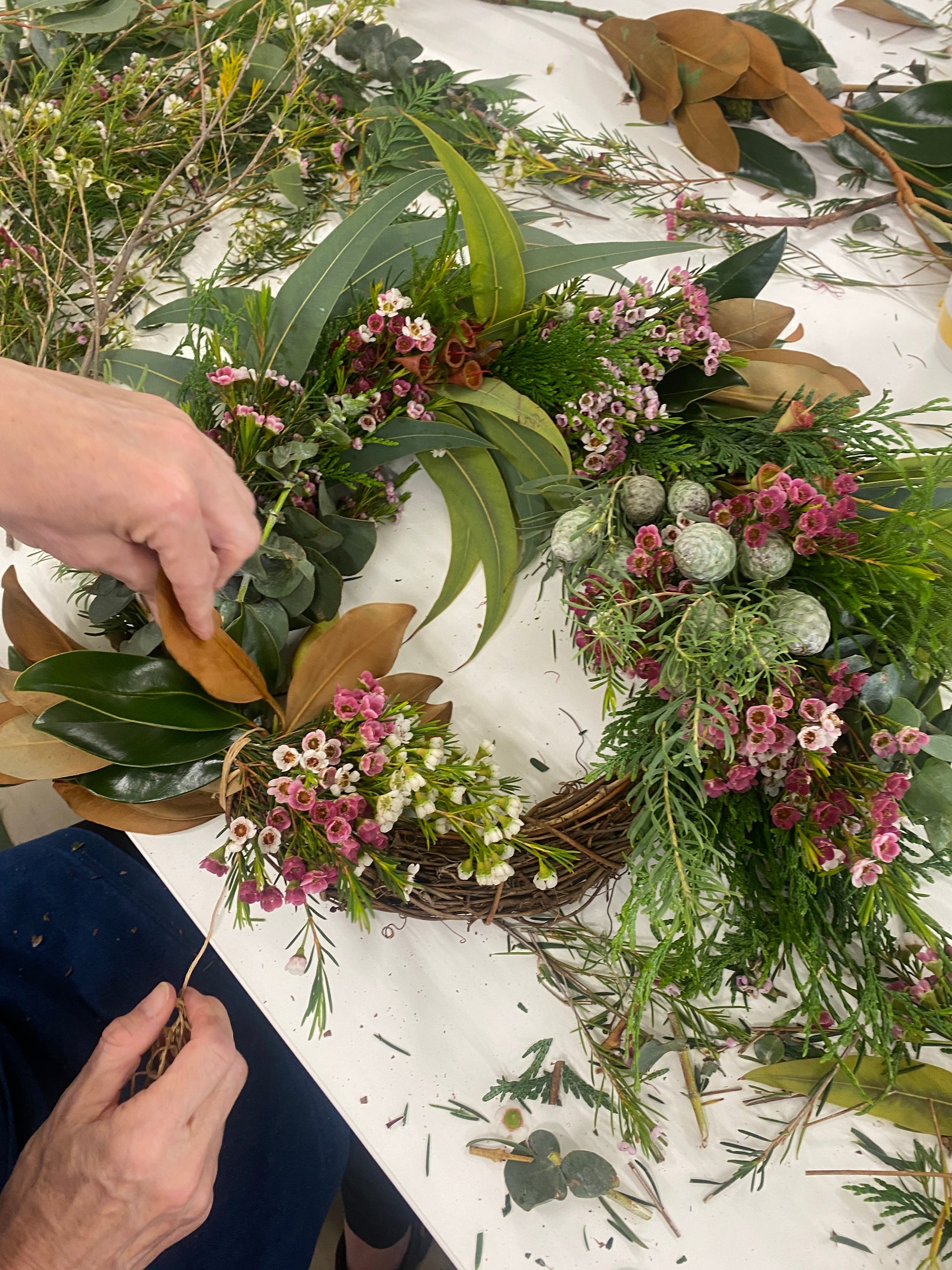 Person crafting a floral wreath with greenery and pink flowers on a white surface.