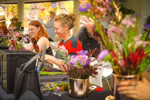 Smiling workshop attendees sitting at tables arranging bright native flowers in metal vases