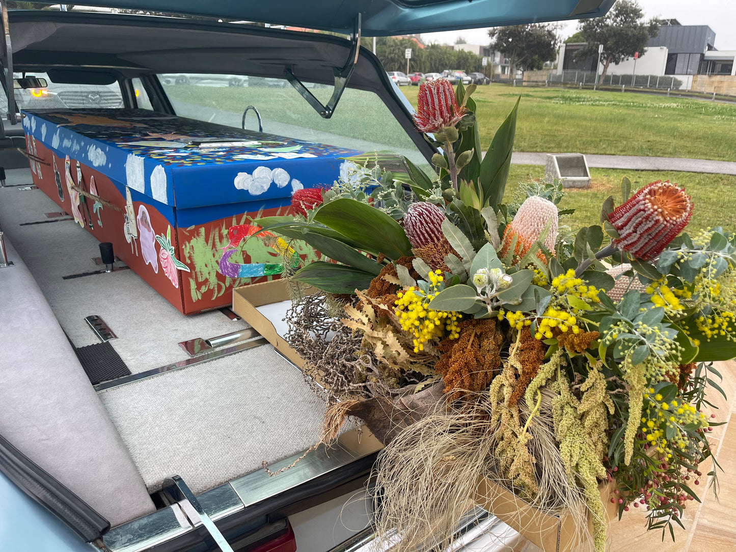 A bespoke Australian native flower  funeral casket flower arrangement next to a hand-painted coffin.